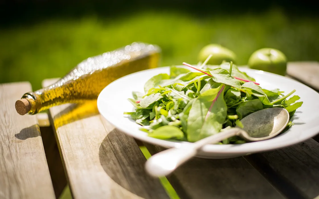 A bowl of green salad on a wooden table beside a bottle of olive oil.