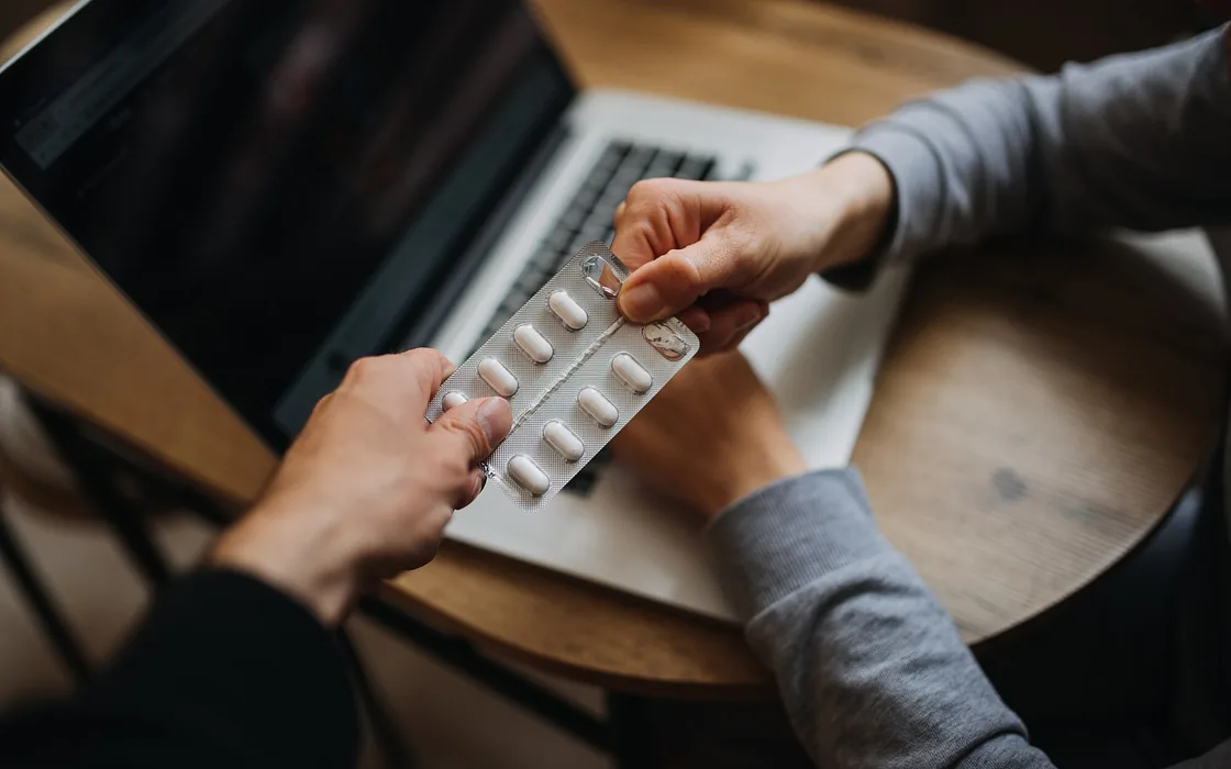Two pairs of hands at a table, one passing a blister pack of tablets to the other.