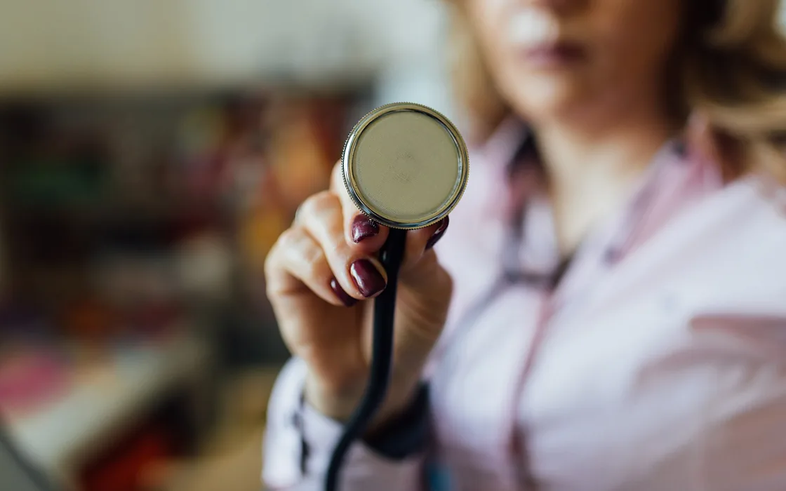 A clinician holding out the chest piece of a stethoscope, the room behind her out of focus.