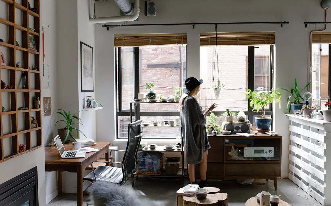 A woman standing at a tall window in a bright room full of plants and books.